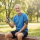 A smiling senior man sits outdoors, using a hand exerciser and listening to music, likely engaging in a workout session.